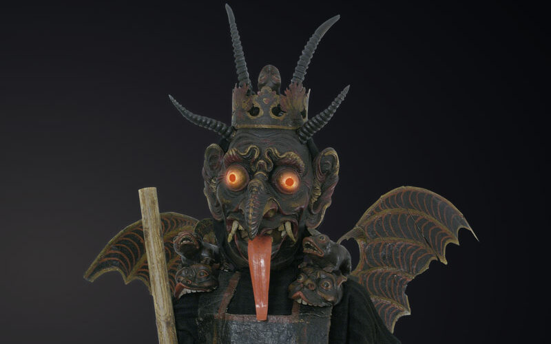 Eine Figur, die eine gehörnte, dämonische Maske mit glühenden Augen, fledermausähnlichen Flügeln und einer Krone trägt und einen Holzstab in der Hand hält, steht vor einem dunklen Hintergrund.