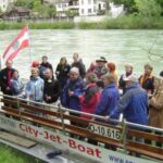 Eine Gruppe von Menschen steht auf einem angedockten Jetboot an einem Fluss und hört einem Mann in einer roten Jacke zu, der ein Mikrofon hält. Am Heck des Bootes ist eine österreichische Flagge angebracht.