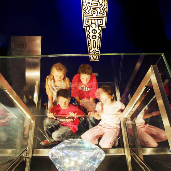 Vier Kinder sitzen auf einer Glasplattform über einer großen beleuchteten Diamantanzeige, an der Wand hinter ihnen ist ein dekoratives Symbol angebracht.