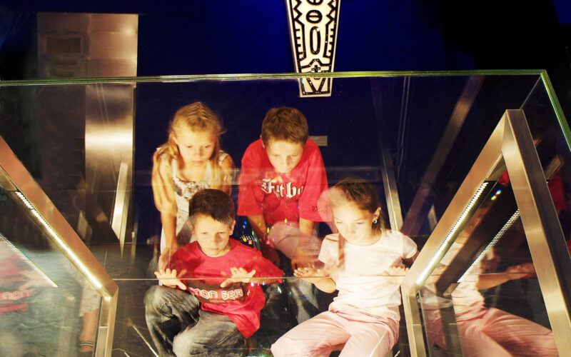 Vier Kinder sitzen auf einer Glasplattform über einer großen beleuchteten Diamantanzeige, an der Wand hinter ihnen ist ein dekoratives Symbol angebracht.