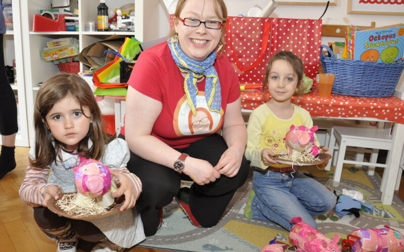 Ein Erwachsener und zwei kleine Kinder sitzen in einem farbenfrohen Raum auf dem Boden und halten rosafarbene Bastelprojekte in der Hand, umgeben von Kunstzubehör.