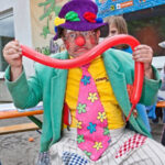 Ein Clown in einem farbenfrohen Outfit und mit Hut hält einen langen roten Luftballon und macht ein albernes Gesicht bei einer Veranstaltung im Freien.