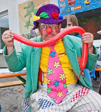 Ein Clown in einem farbenfrohen Outfit und mit Hut hält einen langen roten Luftballon und macht ein albernes Gesicht bei einer Veranstaltung im Freien.