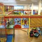 Indoor-Spielplatz mit bunten Klettergerüsten, Rutschen, Ballgruben und spielenden Kindern; einige Erwachsene beaufsichtigen den Autoscooter.