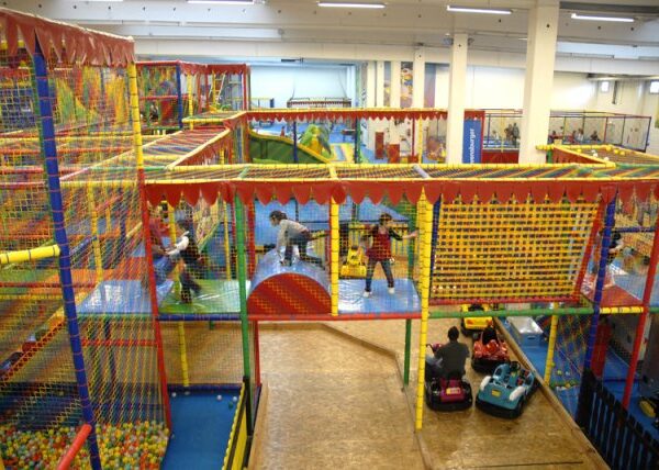 Indoor-Spielplatz mit bunten Klettergerüsten, Rutschen, Ballgruben und spielenden Kindern; einige Erwachsene beaufsichtigen den Autoscooter.