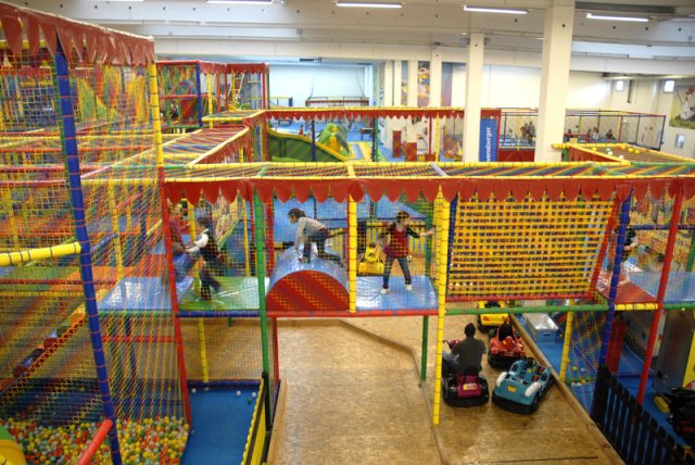 Indoor-Spielplatz mit bunten Klettergerüsten, Rutschen, Ballgruben und spielenden Kindern; einige Erwachsene beaufsichtigen den Autoscooter.