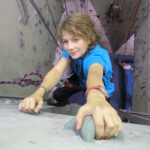 Ein junger Mensch in einem blauen Hemd klettert an einer Indoor-Felswand, hält sich an Griffen fest, während im Hintergrund Seile und ein anderer Kletterer zu sehen sind.