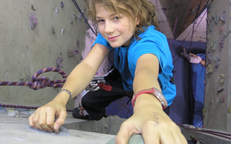 Ein junger Mensch in einem blauen Hemd klettert an einer Indoor-Felswand, hält sich an Griffen fest, während im Hintergrund Seile und ein anderer Kletterer zu sehen sind.