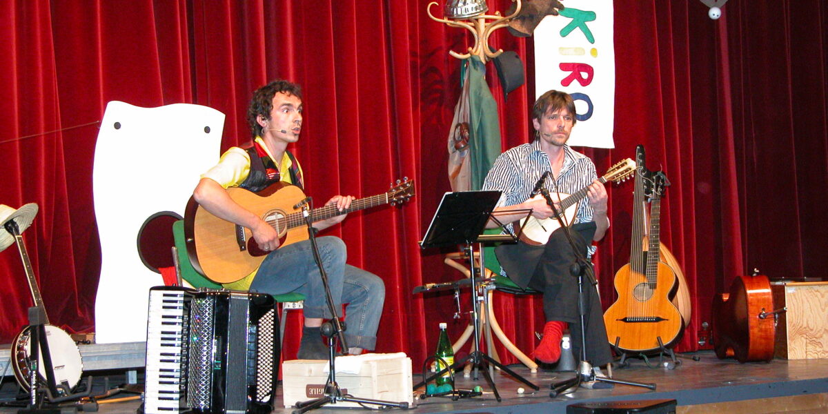 Zwei Musiker sitzen auf der Bühne und spielen Saiteninstrumente, daneben stehen ein Akkordeon und Gitarren. Im Hintergrund sind ein roter Vorhang, Requisiten und ein buntes vertikales Banner zu sehen.