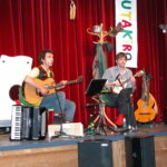 Zwei Musiker sitzen auf der Bühne und spielen Saiteninstrumente, daneben stehen ein Akkordeon und Gitarren. Im Hintergrund sind ein roter Vorhang, Requisiten und ein buntes vertikales Banner zu sehen.