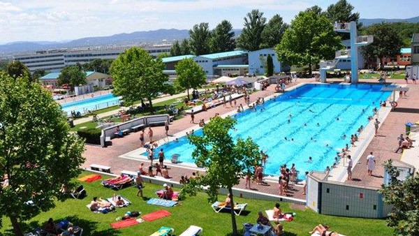Ein großer öffentlicher Freibadkomplex, in dem die Leute schwimmen und andere sich auf der umliegenden Wiese sonnen.