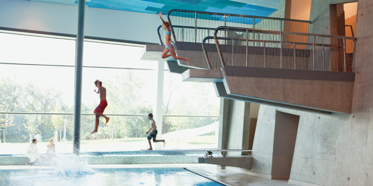 Drei Kinder in Badeanzügen springen von einem Sprungturm in ein Hallenbad, wobei ein Kind bereits ins Wasser platscht.