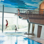 Drei Kinder in Badeanzügen springen von einem Sprungturm in ein Hallenbad, wobei ein Kind bereits ins Wasser platscht.