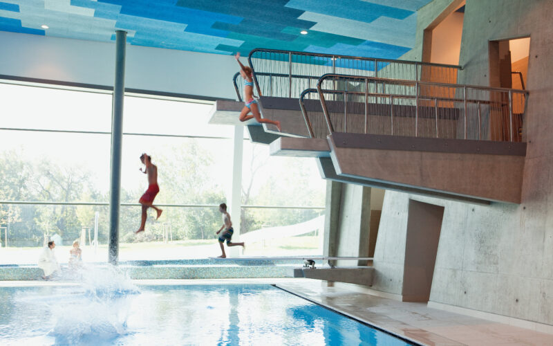 Drei Kinder in Badeanzügen springen von einem Sprungturm in ein Hallenbad, wobei ein Kind bereits ins Wasser platscht.