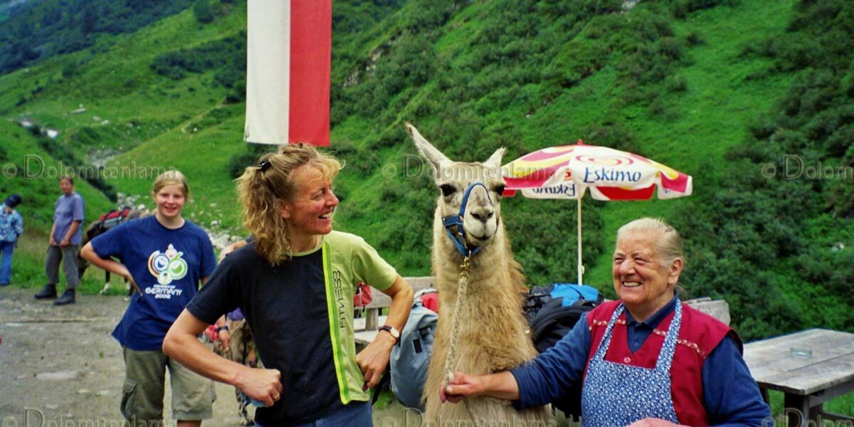 Zwei Menschen stehen lächelnd neben einem Lama auf einem Bergpfad; einer hält die Leine des Lamas. Im Hintergrund sind grüne Hügel und Flaggen zu sehen.
