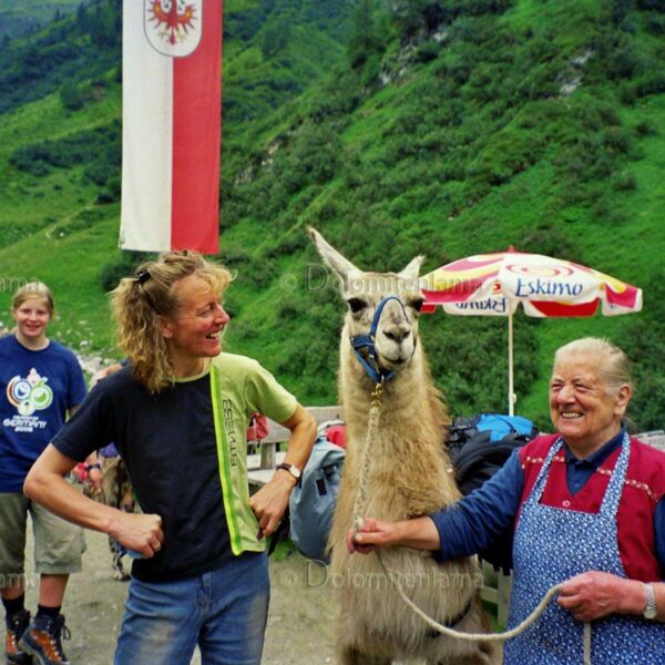 Zwei Menschen stehen lächelnd neben einem Lama auf einem Bergpfad; einer hält die Leine des Lamas. Im Hintergrund sind grüne Hügel und Flaggen zu sehen.