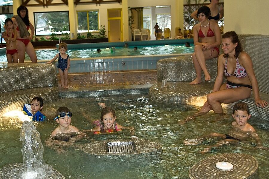 Mehrere Kinder sitzen und spielen in einem flachen Hallenbad, während ein paar Erwachsene und Kinder vom Beckenrand aus zusehen.
