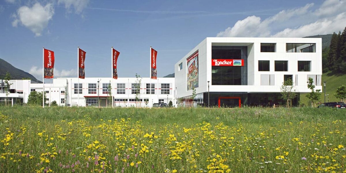 Ein modernes weißes Gebäude mit roten Loacker-Schildern steht hinter einem Feld mit Wildblumen unter einem teilweise bewölkten Himmel.