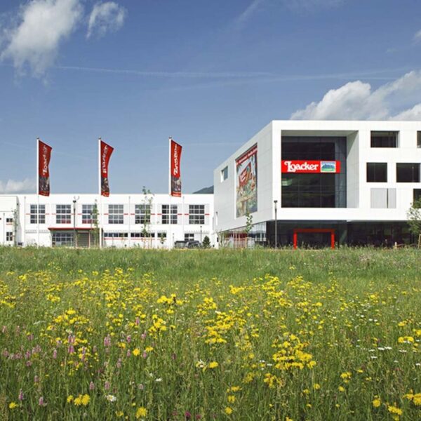 Ein modernes weißes Gebäude mit roten Loacker-Schildern steht hinter einem Feld mit Wildblumen unter einem teilweise bewölkten Himmel.