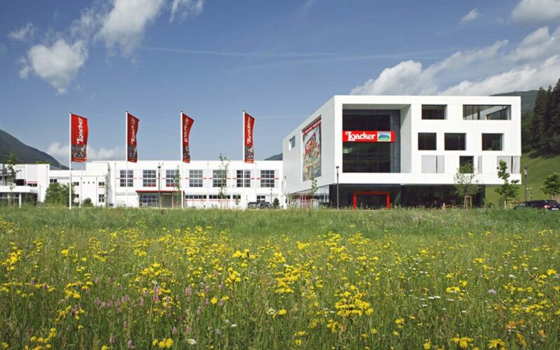 Ein modernes weißes Gebäude mit roten Loacker-Schildern steht hinter einem Feld mit Wildblumen unter einem teilweise bewölkten Himmel.