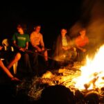 Eine Gruppe junger Leute sitzt nachts im Kreis um ein Lagerfeuer, beleuchtet vom Schein des Feuers. Einige sitzen auf Plastikstühlen, im Feuer sind Holzscheite gestapelt.