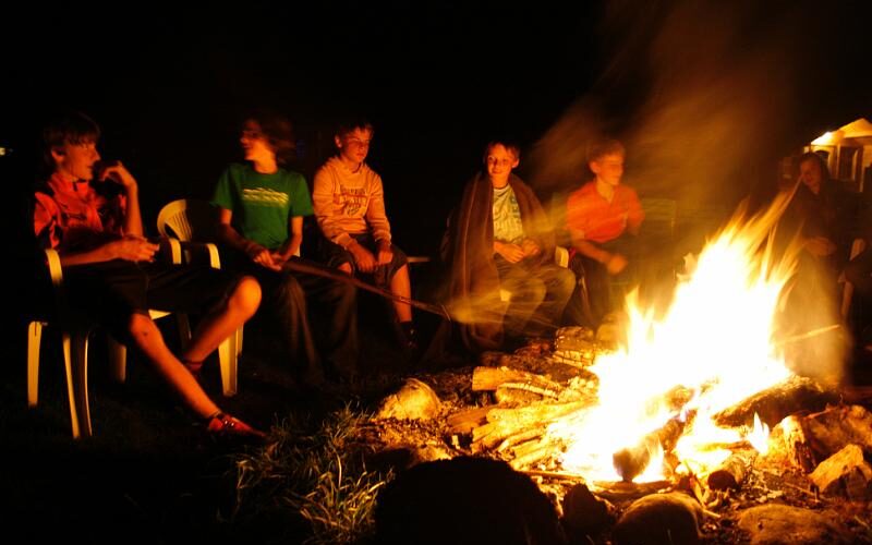 Eine Gruppe junger Leute sitzt nachts im Kreis um ein Lagerfeuer, beleuchtet vom Schein des Feuers. Einige sitzen auf Plastikstühlen, im Feuer sind Holzscheite gestapelt.