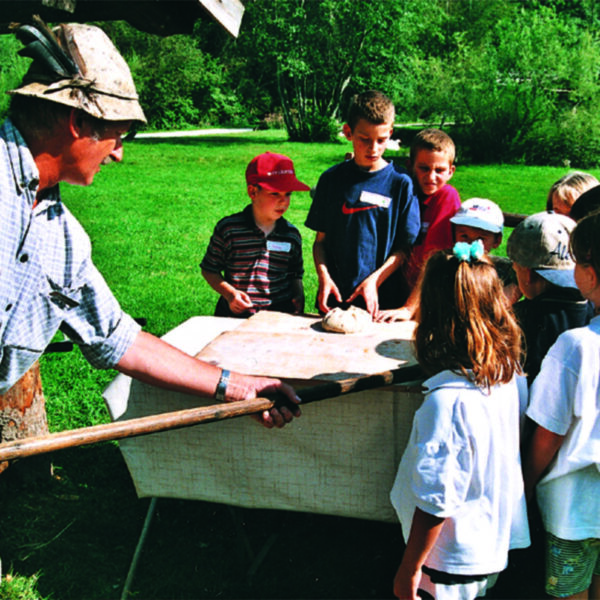 Ein Erwachsener mit Hut demonstriert einer Gruppe von Kindern, die sich im Freien um einen Tisch versammelt haben, das Backen mit einem Holzpaddel. Im Hintergrund sind Bäume und Gras zu sehen.