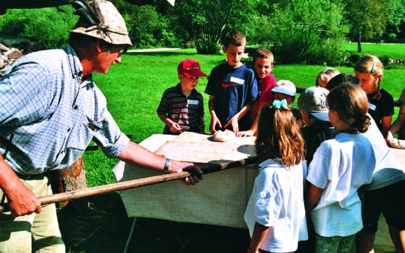Ein Erwachsener mit Hut demonstriert einer Gruppe von Kindern, die sich im Freien um einen Tisch versammelt haben, das Backen mit einem Holzpaddel. Im Hintergrund sind Bäume und Gras zu sehen.