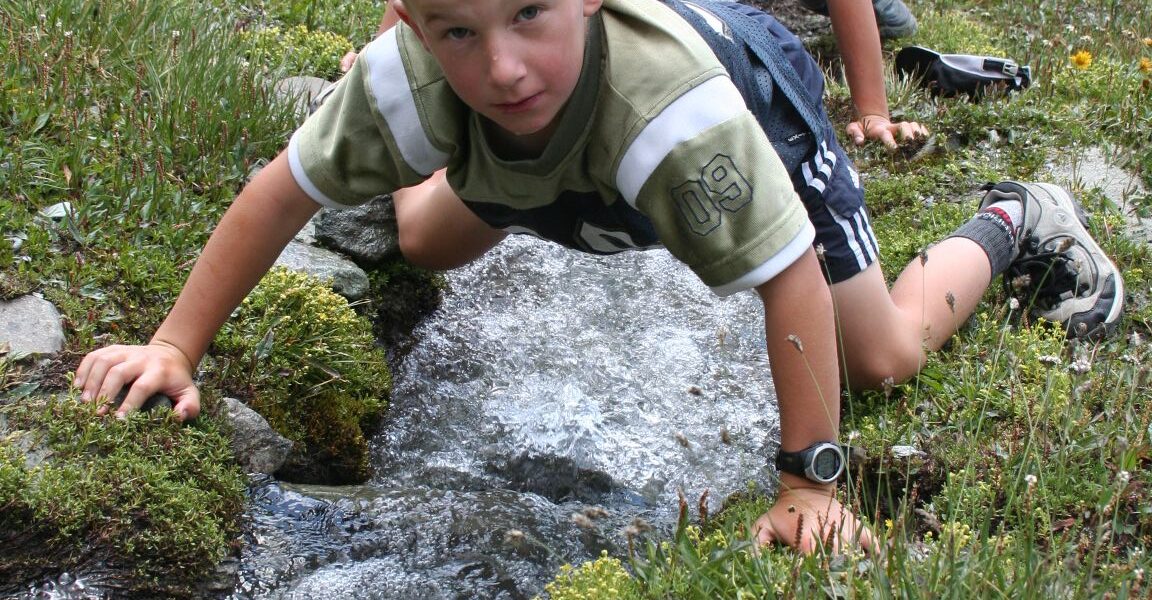 Zwei Jungen knien neben einem kleinen Gebirgsbach, berühren das Wasser und blicken hinunter, umgeben von Gras und Felsen.