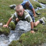Zwei Jungen knien neben einem kleinen Gebirgsbach, berühren das Wasser und blicken hinunter, umgeben von Gras und Felsen.