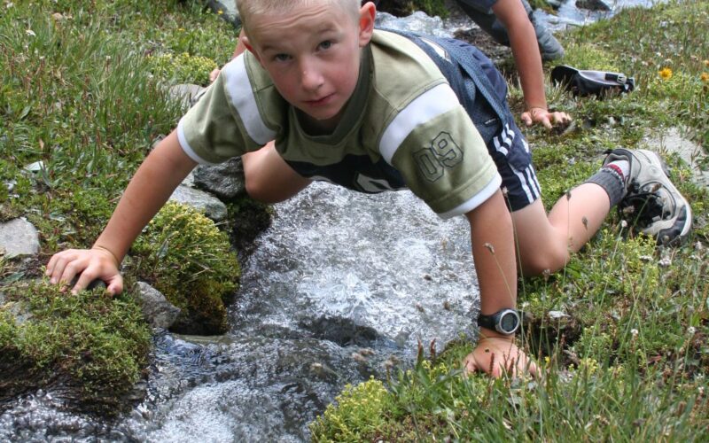 Zwei Jungen knien neben einem kleinen Gebirgsbach, berühren das Wasser und blicken hinunter, umgeben von Gras und Felsen.