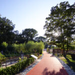 An einem sonnigen Tag führt ein roter Wanderweg durch einen Landschaftspark mit Gartenbeeten, Bäumen und Hinweisschildern.