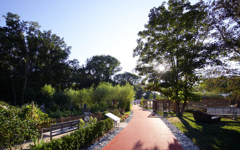 An einem sonnigen Tag führt ein roter Wanderweg durch einen Landschaftspark mit Gartenbeeten, Bäumen und Hinweisschildern.