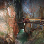 Eine Holzbrücke führt über eine enge, felsige Schlucht mit fließendem Wasser am Grund; steile Felswände umgeben den Weg.