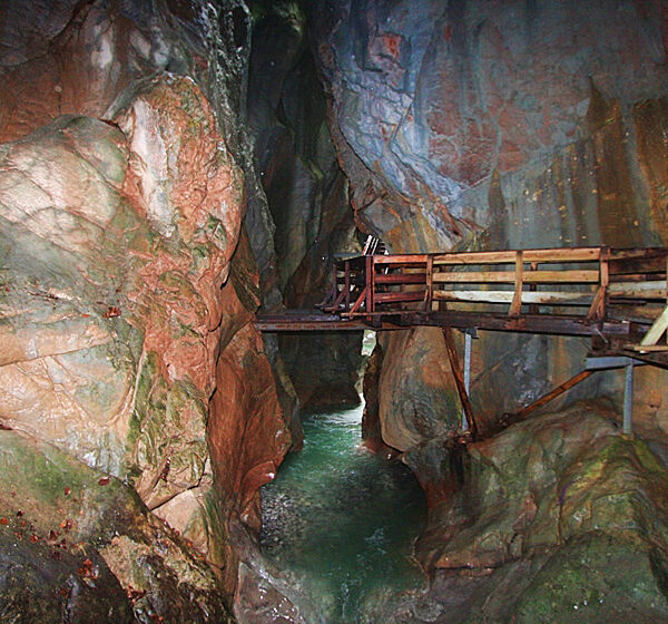 Eine Holzbrücke führt über eine enge, felsige Schlucht mit fließendem Wasser am Grund; steile Felswände umgeben den Weg.