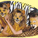 Drei Kinder in Löwenkostümen mit aufgemalten Gesichtern und Outfits mit Tiermustern posieren und lächeln vor einem gelben Hintergrund.