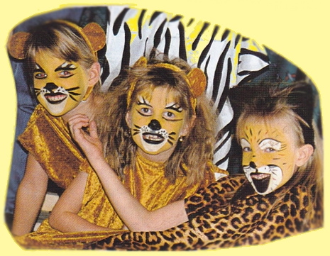 Drei Kinder in Löwenkostümen mit aufgemalten Gesichtern und Outfits mit Tiermustern posieren und lächeln vor einem gelben Hintergrund.