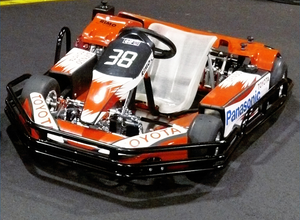 Rot-weißes Go-Kart mit der Nummer 38 auf der Vorderseite, mit Toyota- und Panasonic-Logos, geparkt in einem Innenraum auf einer schwarzen Fläche.