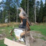 Eine große hölzerne Adlerskulptur steht im Freien auf einem Sockel mit einem Informationsschild und einem schwarzen Stiefel auf der Skulptur. Im Hintergrund sind Bäume und Gras zu sehen.