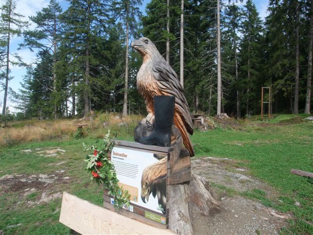 Eine große hölzerne Adlerskulptur steht im Freien auf einem Sockel mit einem Informationsschild und einem schwarzen Stiefel auf der Skulptur. Im Hintergrund sind Bäume und Gras zu sehen.