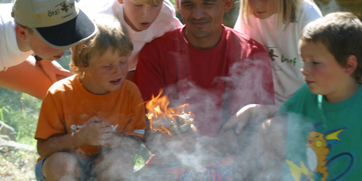 Eine Gruppe von Kindern und ein Erwachsener beobachten aufmerksam, wie ein kleines Feuer auf einem Stück Holz brennt und der Rauch um sie herum aufsteigt.