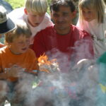 Eine Gruppe von Kindern und ein Erwachsener beobachten aufmerksam, wie ein kleines Feuer auf einem Stück Holz brennt und der Rauch um sie herum aufsteigt.