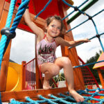 Ein junges Mädchen klettert auf einer blauen Seilbrücke auf einem hölzernen Spielplatz, lächelt und hält sich an den Seilen fest.