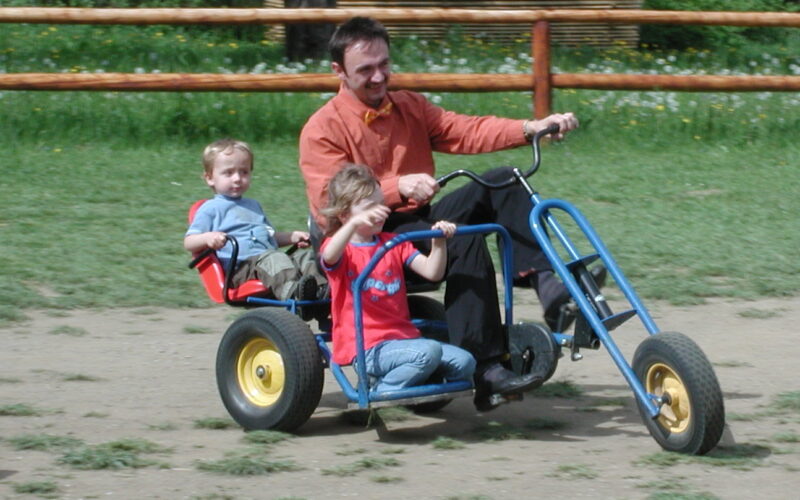 Ein Erwachsener und zwei Kinder fahren auf einem großen Dreirad mit Tretantrieb auf einem unbefestigten Weg in einer Außenanlage mit Gras und Holzzaun.
