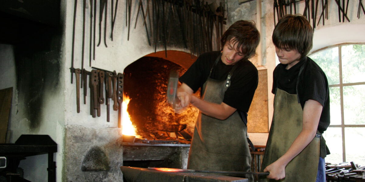 Zwei Personen in Schürzen arbeiten gemeinsam an einem Amboss in einer Schmiedewerkstatt. An der Wand hängen verschiedene Werkzeuge, hinter ihnen glüht in der Esse ein Feuer.