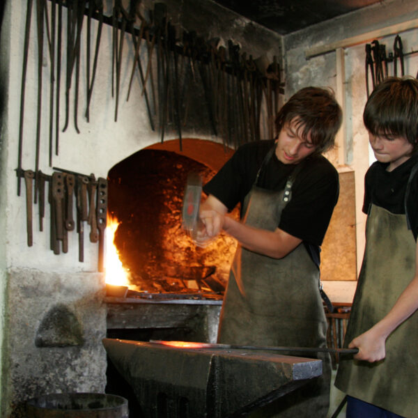 Zwei Personen in Schürzen arbeiten gemeinsam an einem Amboss in einer Schmiedewerkstatt. An der Wand hängen verschiedene Werkzeuge, hinter ihnen glüht in der Esse ein Feuer.