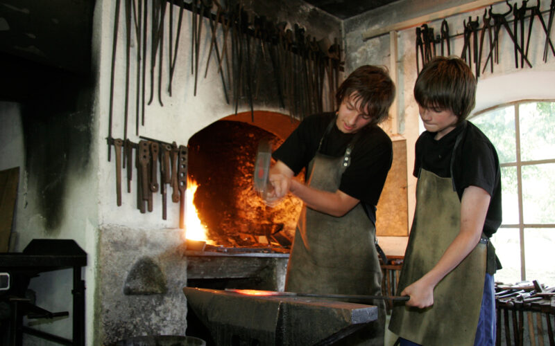Zwei Personen in Schürzen arbeiten gemeinsam an einem Amboss in einer Schmiedewerkstatt. An der Wand hängen verschiedene Werkzeuge, hinter ihnen glüht in der Esse ein Feuer.