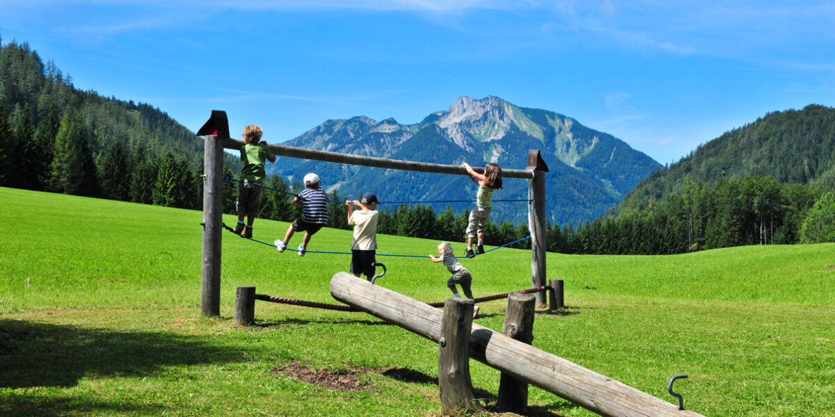 Fünf Kinder spielen auf einem hölzernen Klettergerüst auf einer Wiese mit einem Berg und Bäumen im Hintergrund unter einem blauen Himmel.