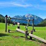Fünf Kinder spielen auf einem hölzernen Klettergerüst auf einer Wiese mit einem Berg und Bäumen im Hintergrund unter einem blauen Himmel.