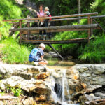 Ein Kind mit Rucksack hockt neben einem kleinen Wasserfall, während ein Erwachsener und ein weiteres Kind auf einer Holzbrücke in einem grünen Waldgebiet stehen.